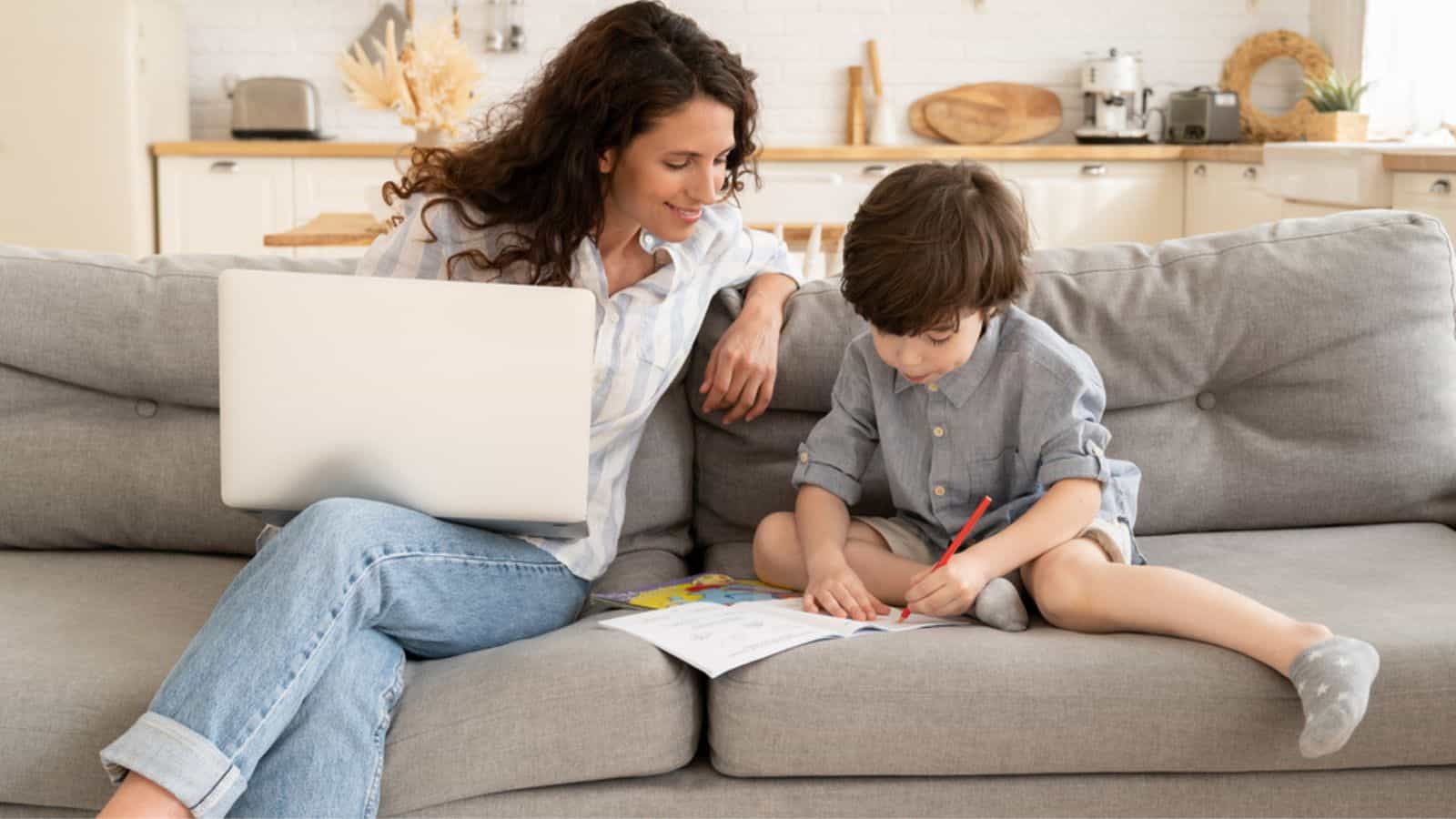 Young mother remote worker businesswoman with preschooler son on couch working on laptop computer