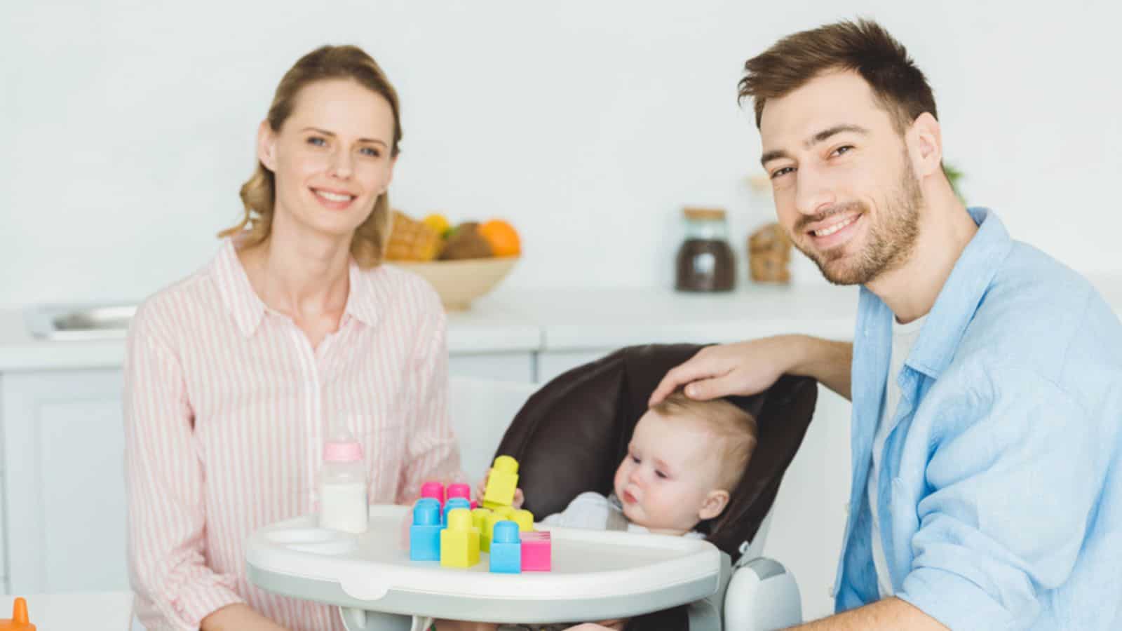 Young parents with infant daughter sitting in baby chair with plastic blocks and feeding bottle