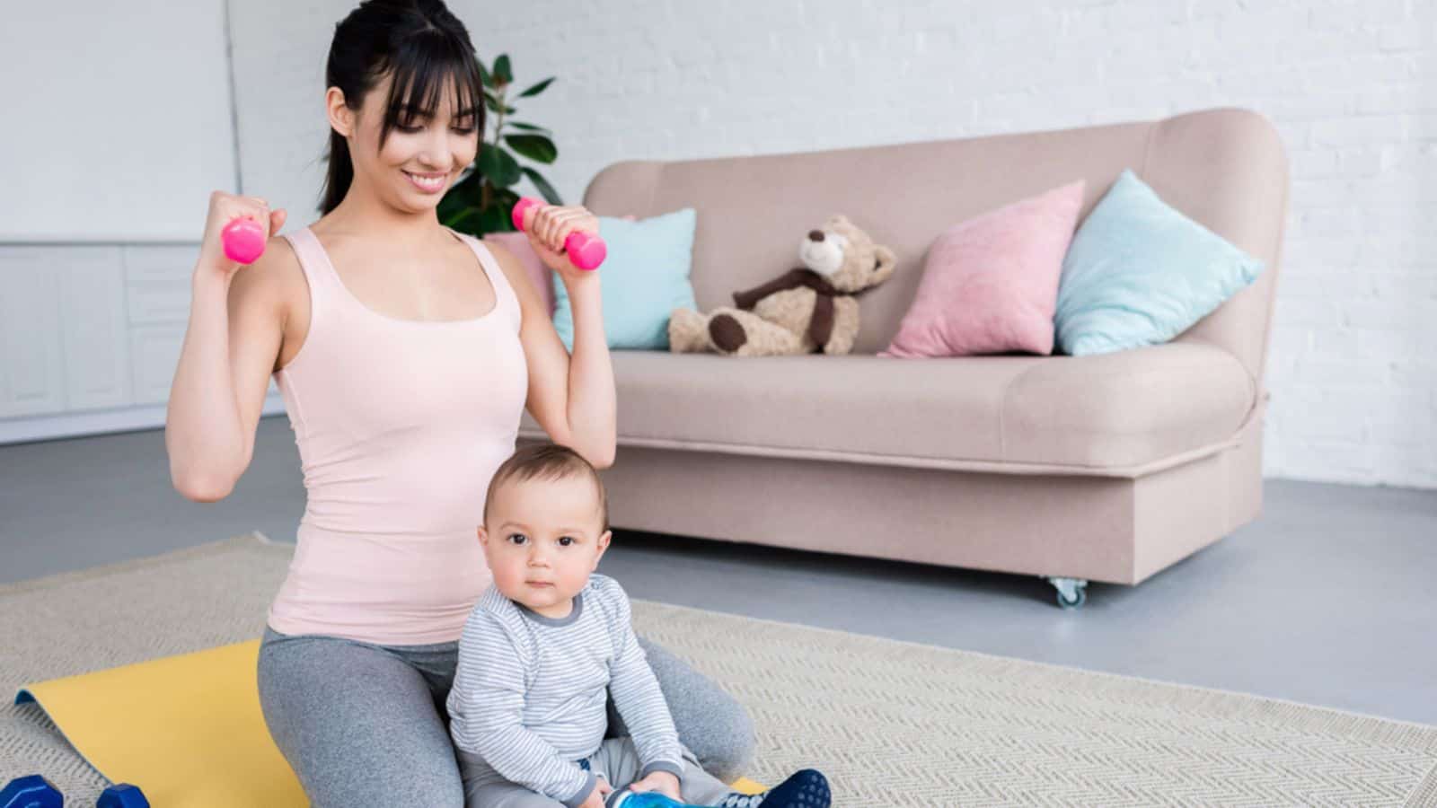 Young smiling mother working out on yoga mat with her little baby