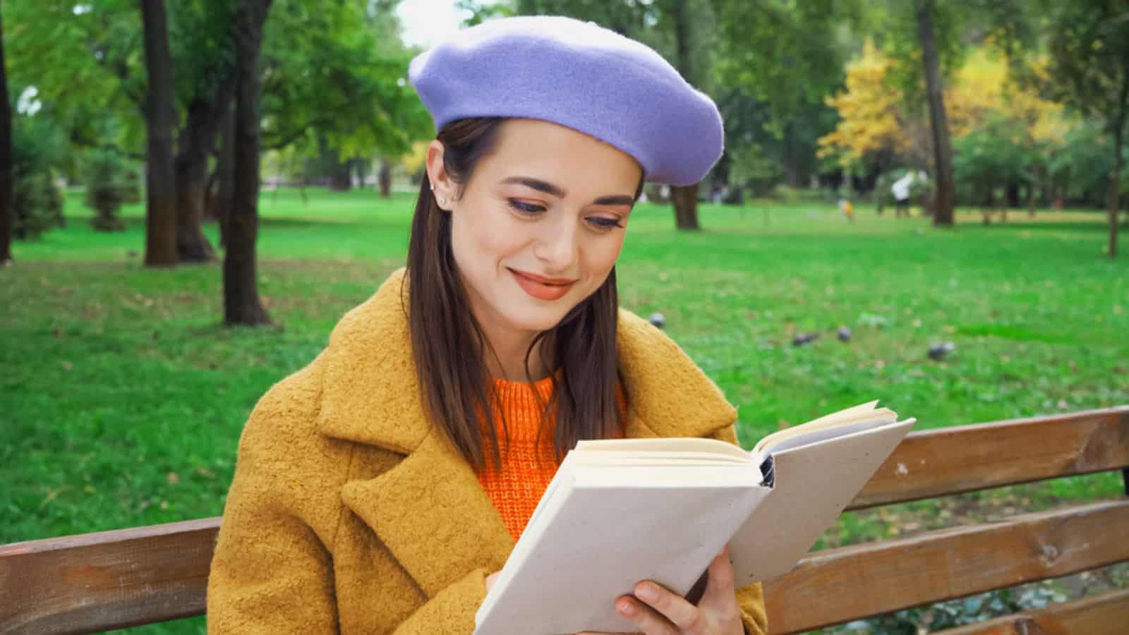 Young stylish woman smiling while reading novel book in autumn park