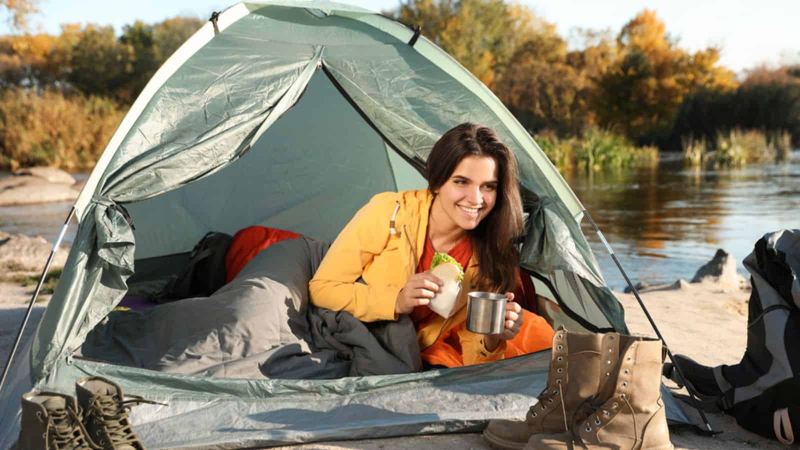 Young woman having breakfast in sleeping bag inside of camping tent