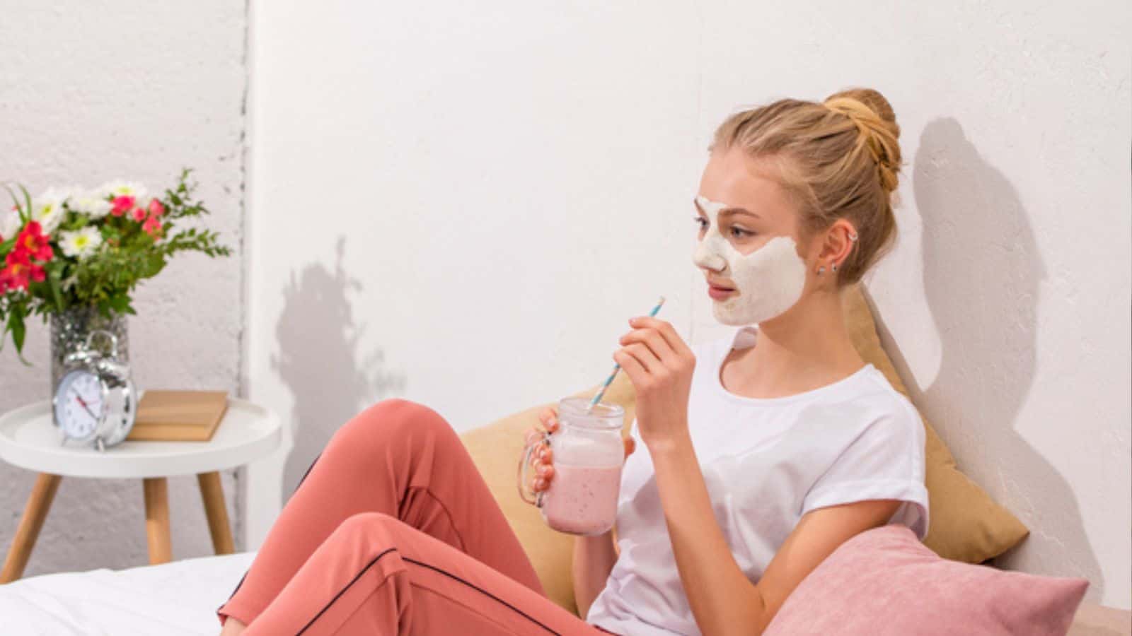 Young woman with white clay facial mask drinking milkshake on bed at home
